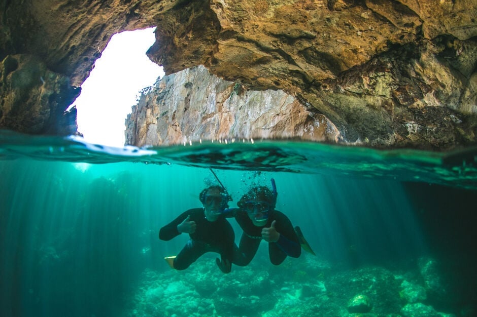 Two scuba divers explore turquoise underwater cave with rocky cliffs visible above water