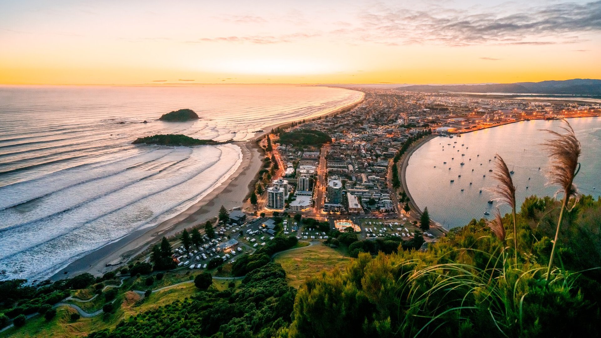 Aerial sunset view of a coastal city with a curved beach, lagoon, and mountains in the distance