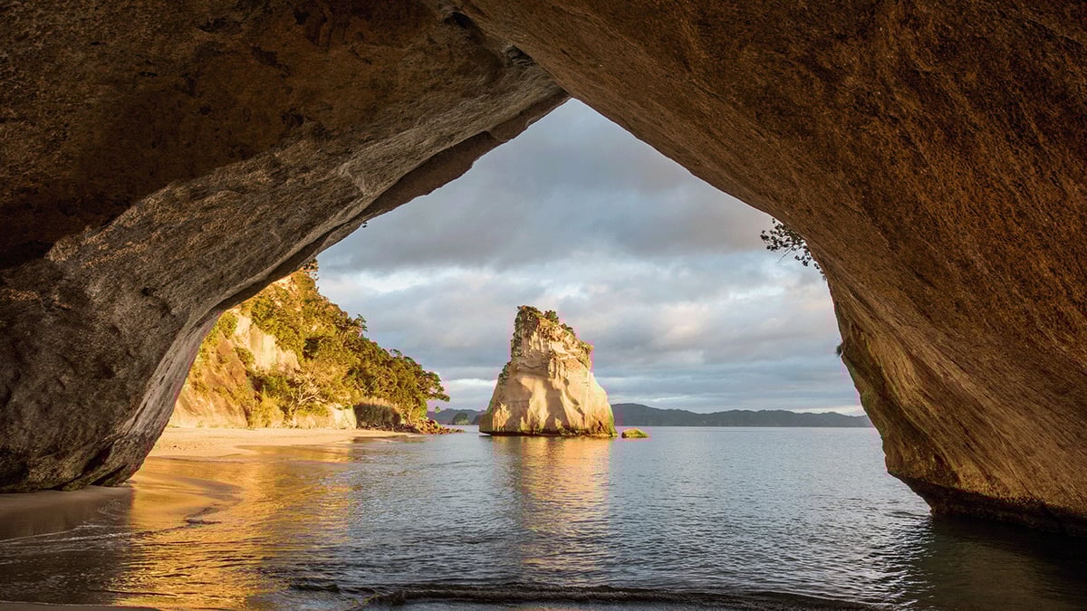 View from inside a sea cave looking out at golden cliffs, a rock formation, and calm ocean waters under cloudy skies