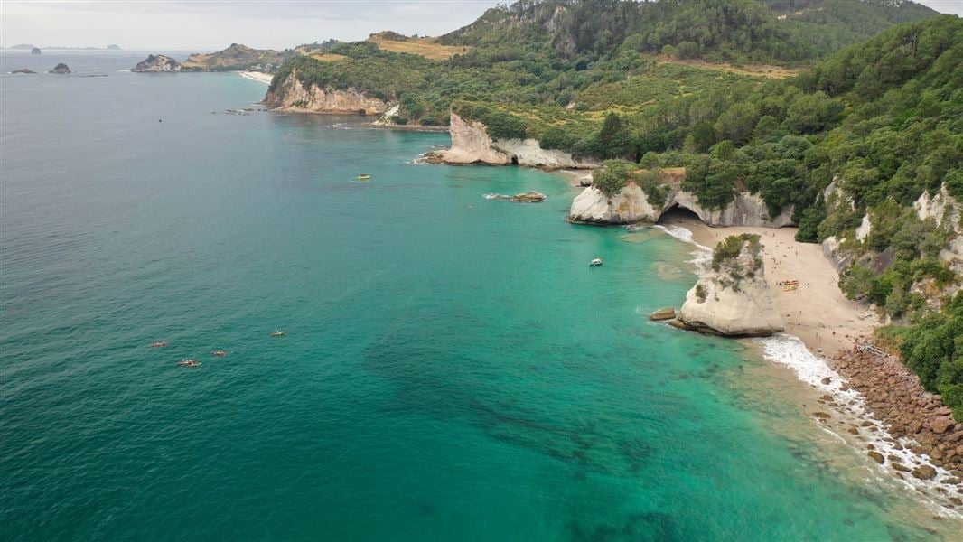 Aerial view of a turquoise cove with white limestone cliffs, green forested hills, and boats anchored in crystal clear Mediterranean waters