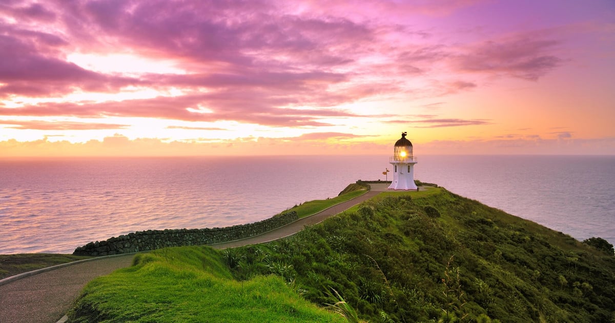 Lighthouse on a green headland at sunset with purple and pink sky over calm ocean waters