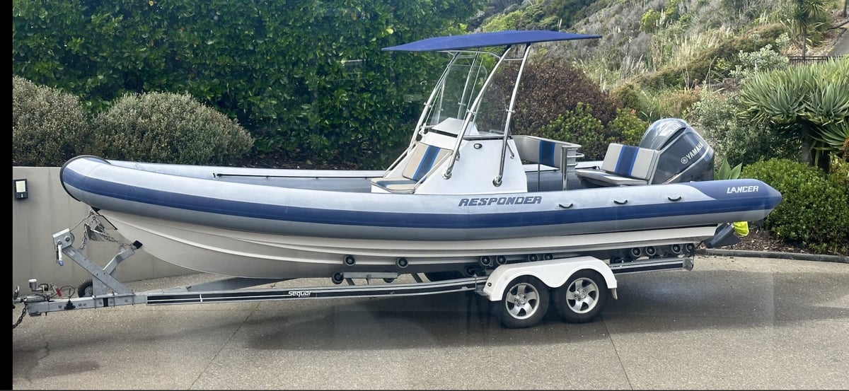 Blue and white rigid inflatable boat with T-top canopy on a dual-axle trailer parked in a driveway