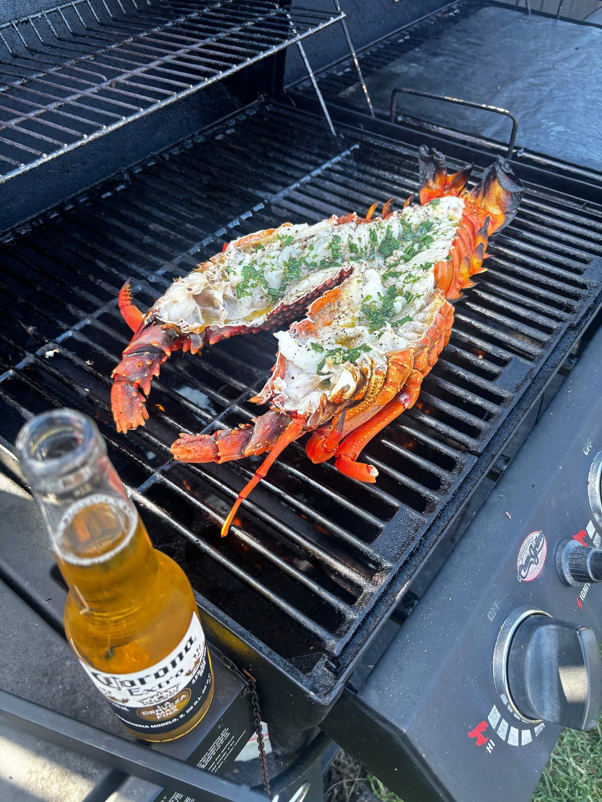 Two grilled lobster halves topped with herbs and butter on a barbecue grill, with a beer bottle beside