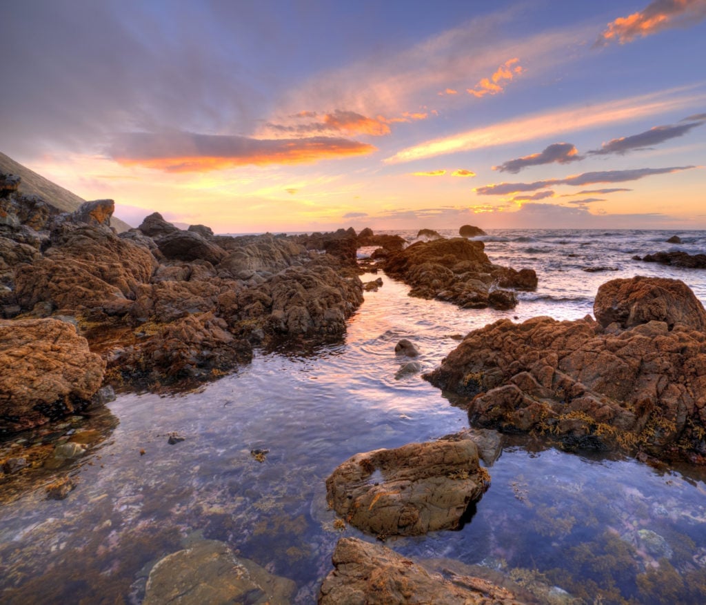 Rocky coastal landscape at sunset with golden sky, calm tidal pools reflecting the colorful clouds