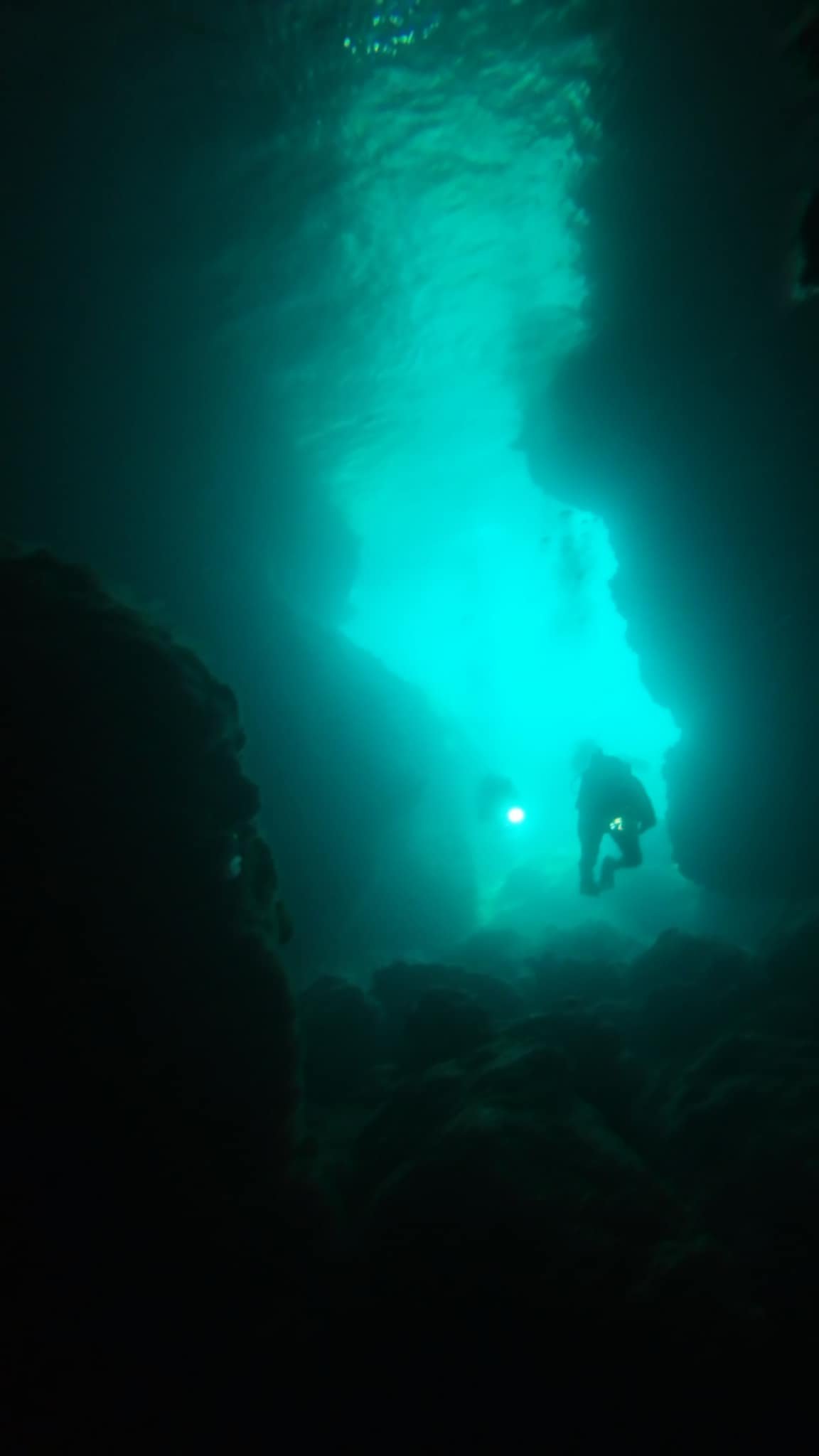 Scuba divers exploring a dark underwater cave with bright turquoise light filtering from above